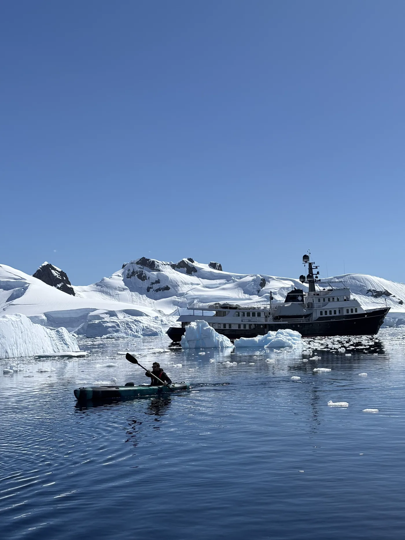 Captain Pavlos kayaking in a glassy Antarctic bay with an explorer yacht at anchor behind